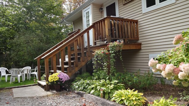 Una casa con escalera de madera que da a un balcón, rodeada por un jardín con diversas plantas y flores.