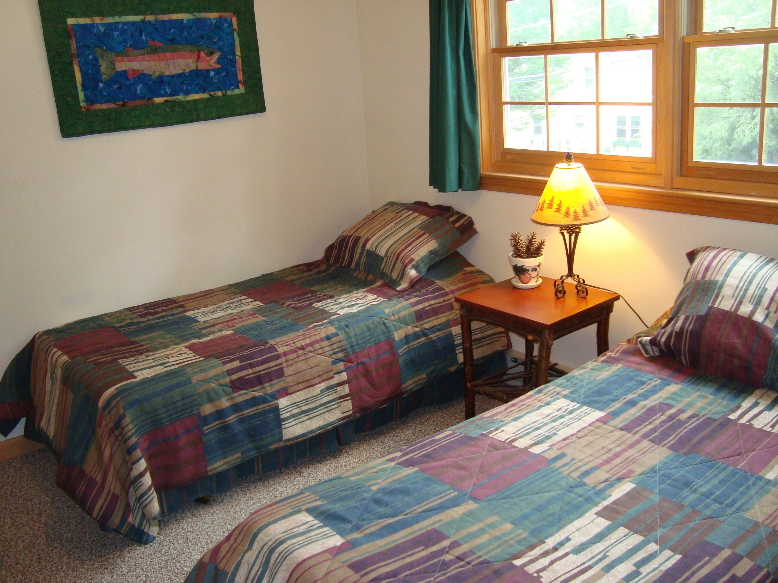 Une chambre avec deux lits simples, une table de chevet avec une lampe, un tableau au mur et une fenêtre avec des rideaux.