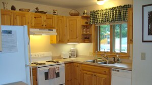 Cocina compacta con armarios de madera, estufa blanca, pileta y ventana con cortina a cuadros.