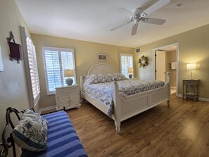 Une chambre à coucher comprenant un lit, un ventilateur de plafond, une fenêtre avec des stores, une table de chevet avec une lampe et une porte.
