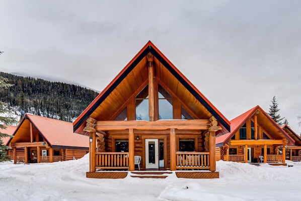 A log cabin with a prominent front porch and large windows, surrounded by snow.