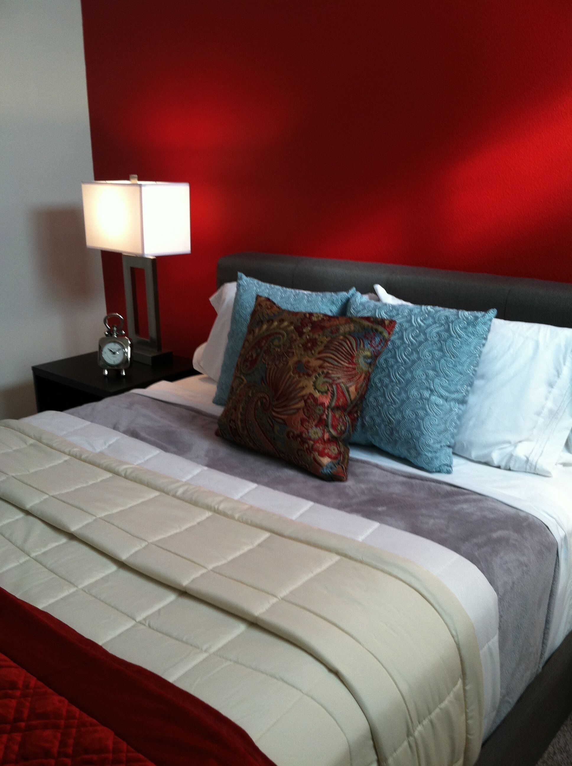 Un lit bien fait, recouvert d’une courtepointe matelassée, un mur rouge et une table de chevet avec une lampe et un réveil.