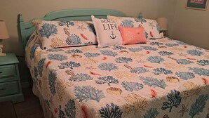 Une chambre à coucher avec un lit recouvert d’une courtepointe à motifs floraux, un tableau représentant une scène de plage accroché au mur et une tête de lit de couleur turquoise.