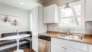 A modern kitchen with white cabinetry, a stainless steel dishwasher, a granite countertop, and a window with a view of trees. - Charming Stones Throw Cottage walk to UNT! (Denton)