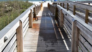 En träpromenad som leder till en strand med en flagga, gräsytor och en klar himmel.