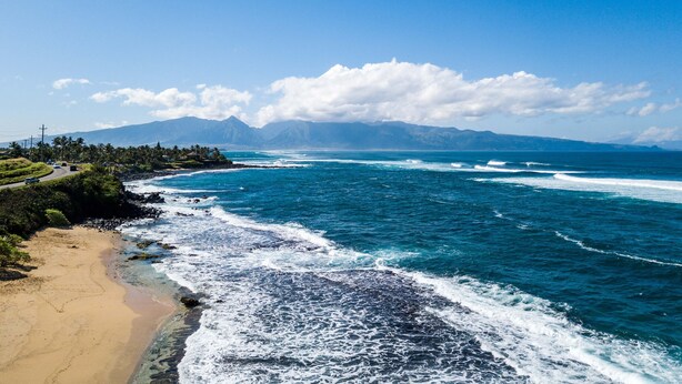 A coastal landscape with a beach, ocean waves, and a mountain range in the background.