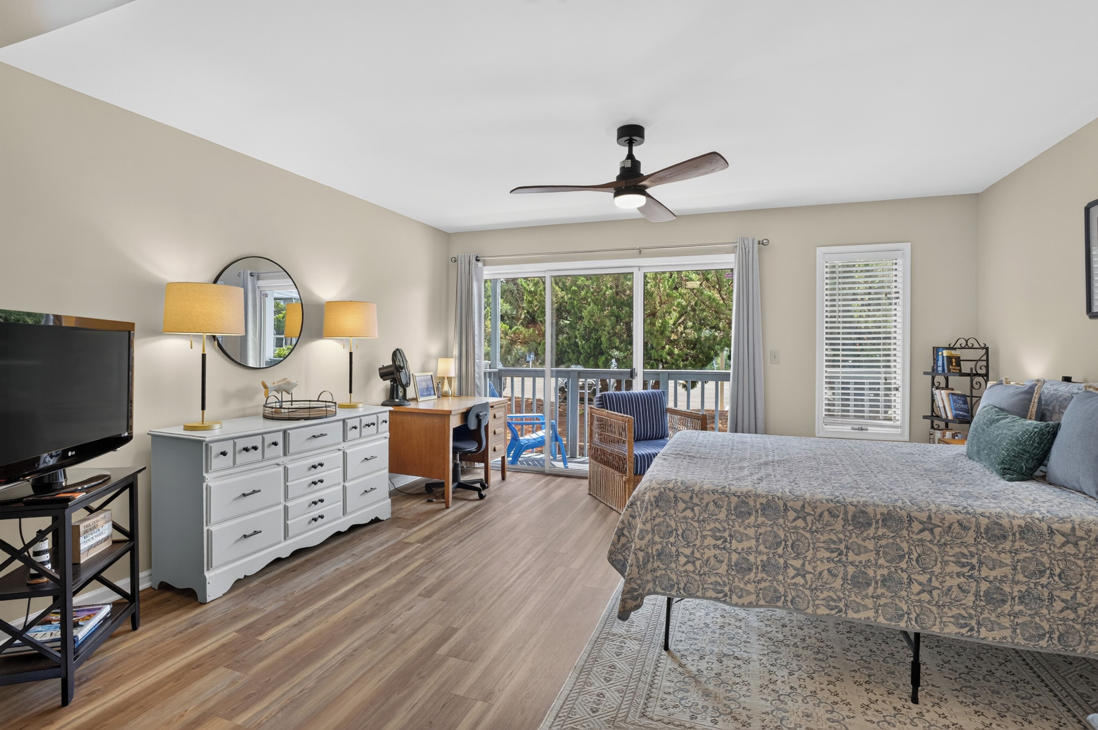 A bedroom with a bed, a desk, a television, a balcony, and a ceiling fan.