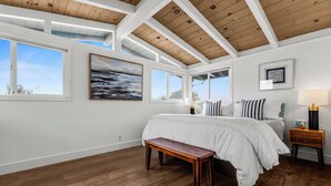 Une chambre avec un grand lit, un plancher en bois, des murs blancs et un plafond en bois. On y trouve des fenêtres, un banc et une table de chevet avec une lampe.