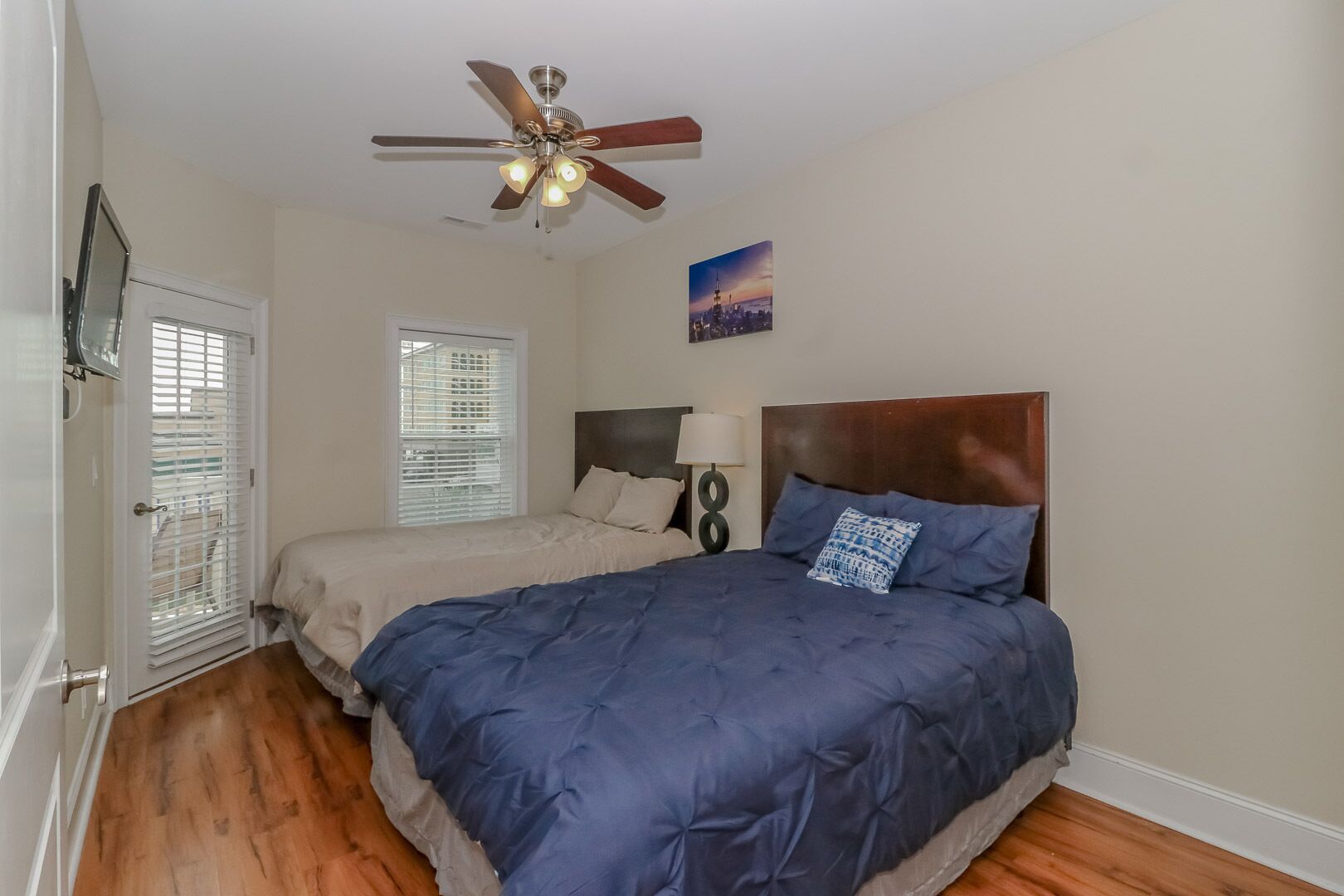 A bedroom with a bed, a ceiling fan, a window with blinds, a wall-mounted lamp, and a cityscape painting.