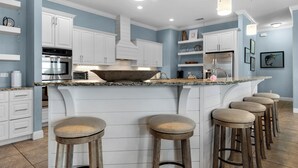 A modern kitchen with white cabinetry, a large island with stools, and pendant lighting. - ☀️ The Beach House | Designer 7BR w/ Huge Pool, Bunk Room + In-Law Suite (Pensacola Beach)
