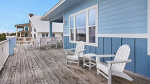 A wooden deck with white Adirondack chairs and a blue building with large windows. - ☀️ The Beach House | Designer 7BR w/ Huge Pool, Bunk Room + In-Law Suite (Pensacola Beach)