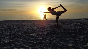 Une plage où une personne pratique une posture de yoga, une autre personne se trouve à l’arrière-plan et le soleil se couche sur l’océan.