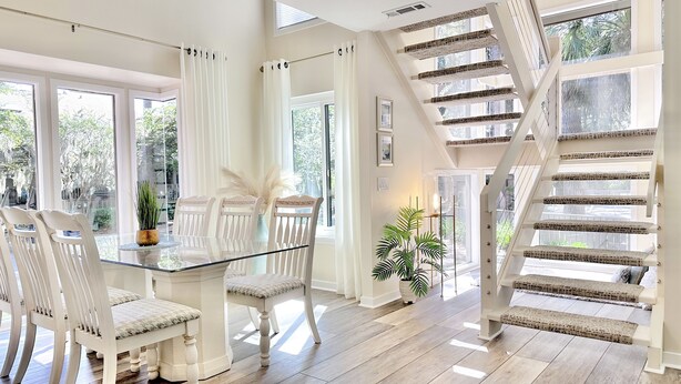 Una habitación amplia con una mesa de comedor de vidrio, sillas blancas, una escalera con escalones estampados y grandes ventanales que dan a vistas de vegetación.