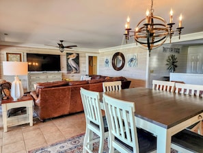 A spacious living room with a brown leather sectional sofa, a dining table with white chairs, a chandelier, and a flat-screen TV mounted on the wall. - Views..Views..Views! Sleeps 21 ! 8 Bedrooms plus Amenity Cards! (Fripp Island)