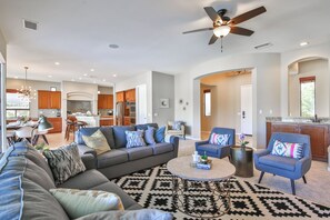 A spacious living room with a gray sectional sofa, a coffee table, and a patterned rug. There is a ceiling fan, recessed lighting, and a view into the kitchen area.