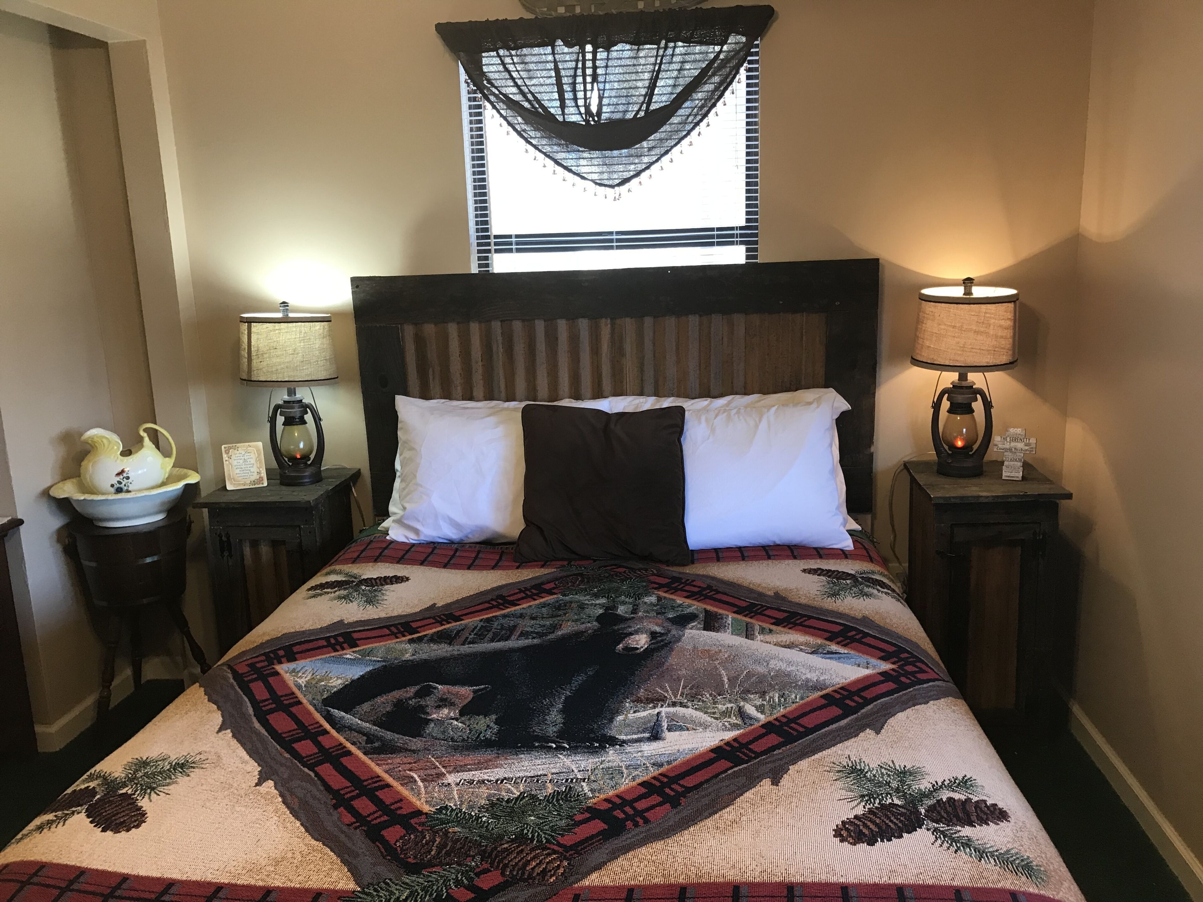 Una cama con colcha que muestra un oso y pinos, flanqueada por dos mesitas de noche con lámparas y un cuenco decorativo.