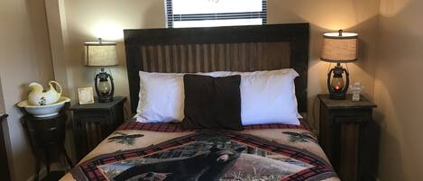 Una cama con colcha que muestra un oso y pinos, flanqueada por dos mesitas de noche con lámparas y un cuenco decorativo.
