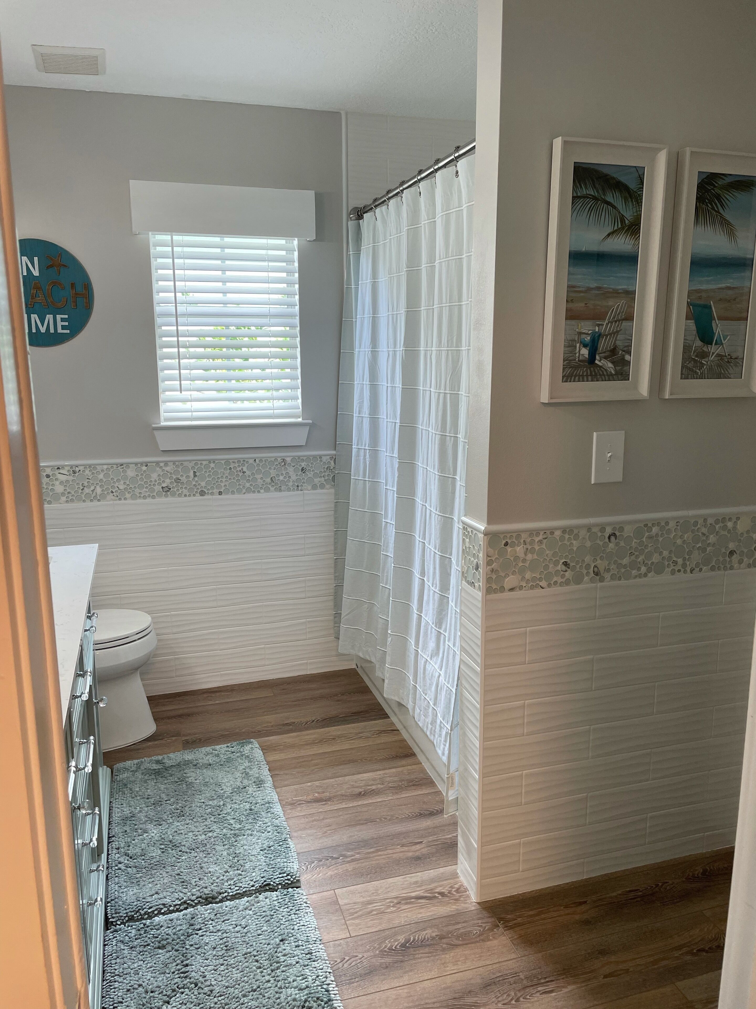 Un baño con inodoro, cortina de ducha, ventana con persianas e imagen de playa en la pared.