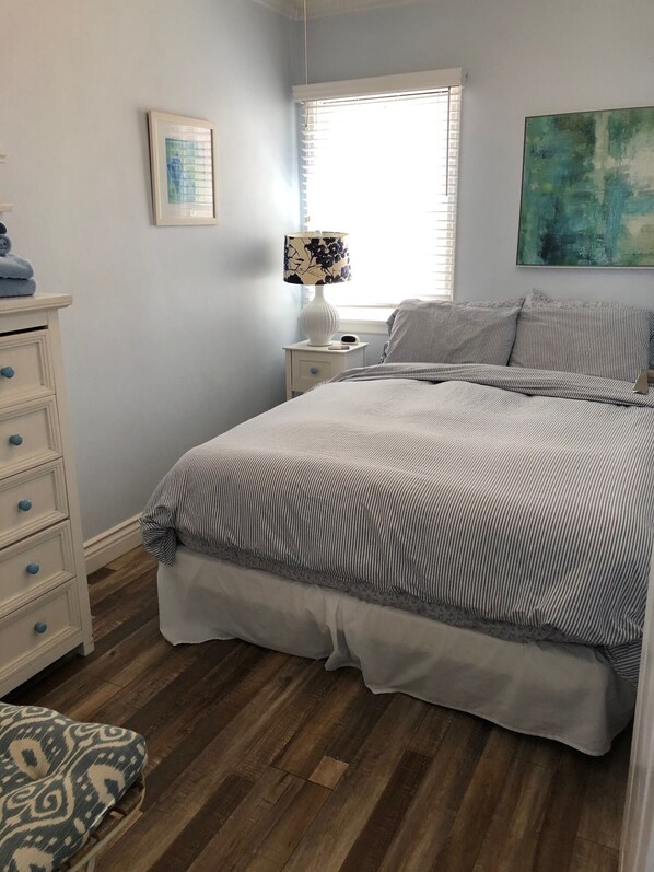 Un dormitorio con cama, mesita de noche, lámpara, cómoda, ventana y un cuadro.