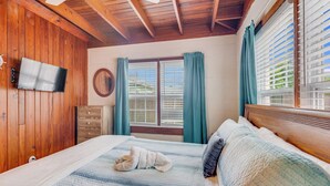 Une chambre avec un plafond en bois, un lit recouvert d’une courtepointe, une commode, un téléviseur fixé au mur et une fenêtre avec des rideaux.