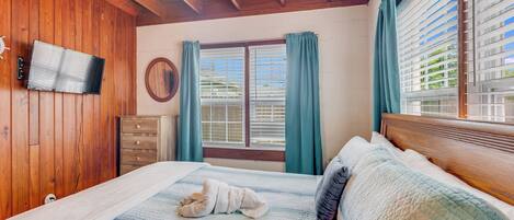 Un dormitorio con techo de madera, una cama con colcha, un aparador, un televisor en la pared y una ventana con cortinas.