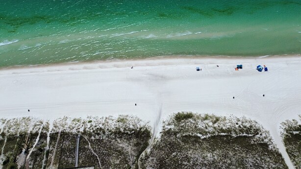 Vue aérienne d’une plage aux eaux turquoise et limpides, bordée d’un rivage sablonneux et d’une végétation clairsemée.