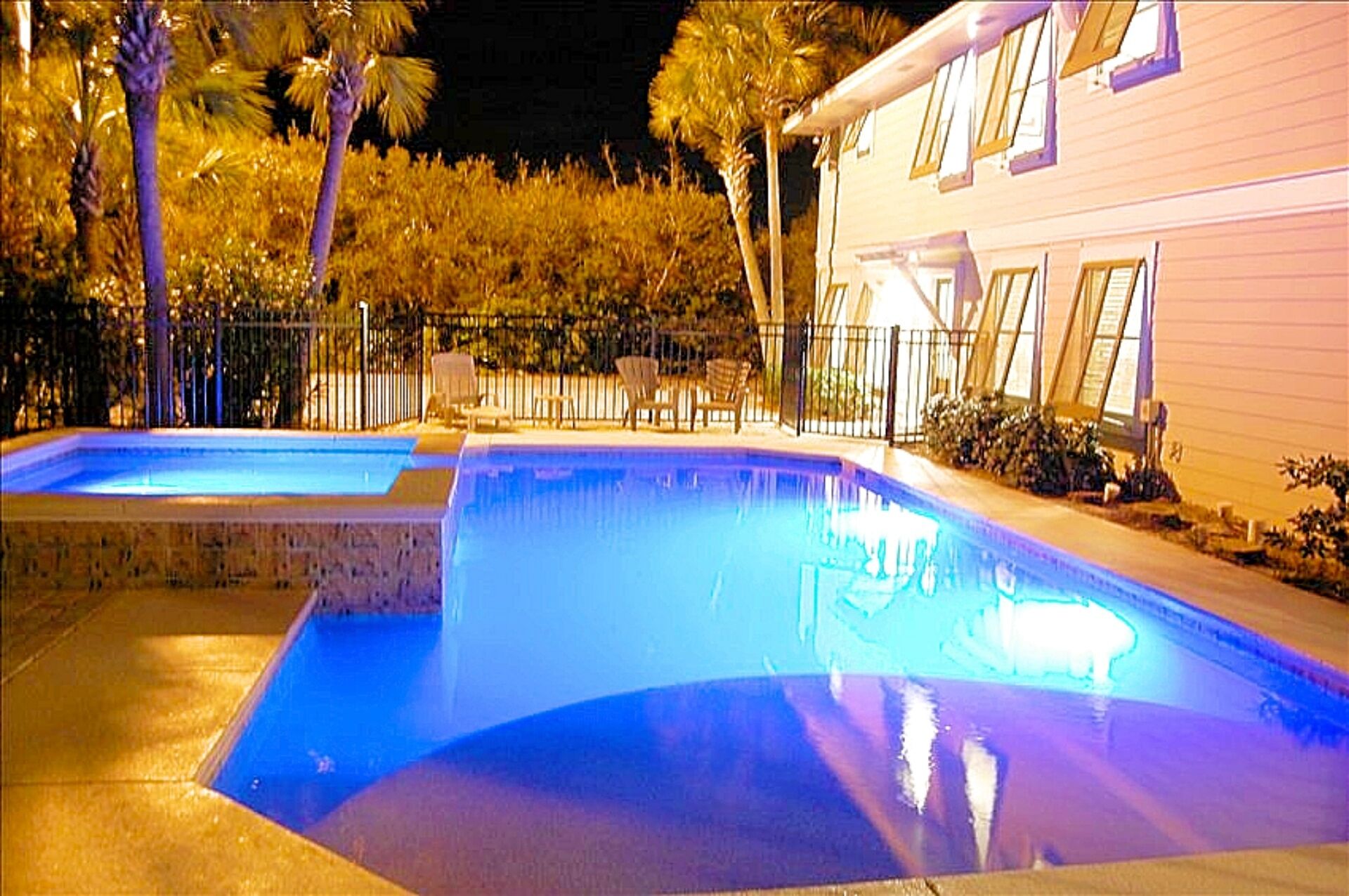 Zona de piscina con dos piscinas, rodeada por una valla, palmeras y un edificio con ventanas.