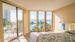 Habitación de hotel con balcón grande, cama con colcha estampada y vista a un resort con piscina y palmeras.