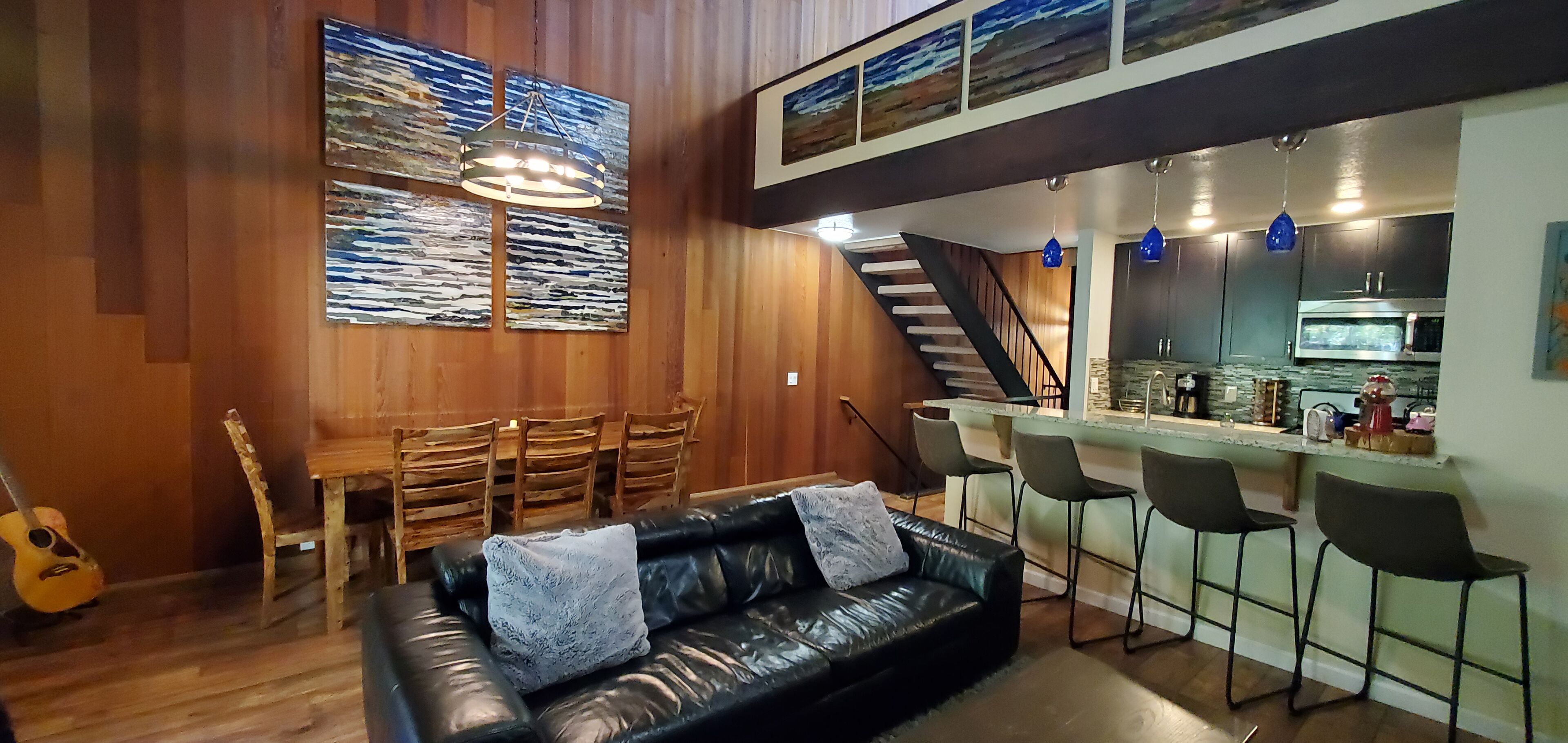 A cozy living area with a black leather sofa, wooden dining table, and chairs. A staircase leads to a kitchen area with a bar and stools.