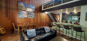 A cozy living area with a black leather sofa, wooden dining table, and chairs. A staircase leads to a kitchen area with a bar and stools.