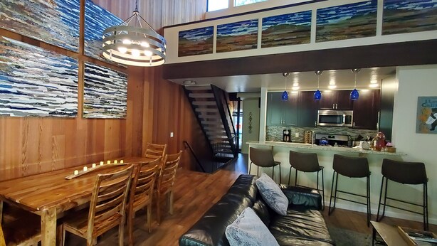 Zona pranzo con tavolo e sedie in legno, un bancone con sgabelli e una scala che conduce a un livello superiore.