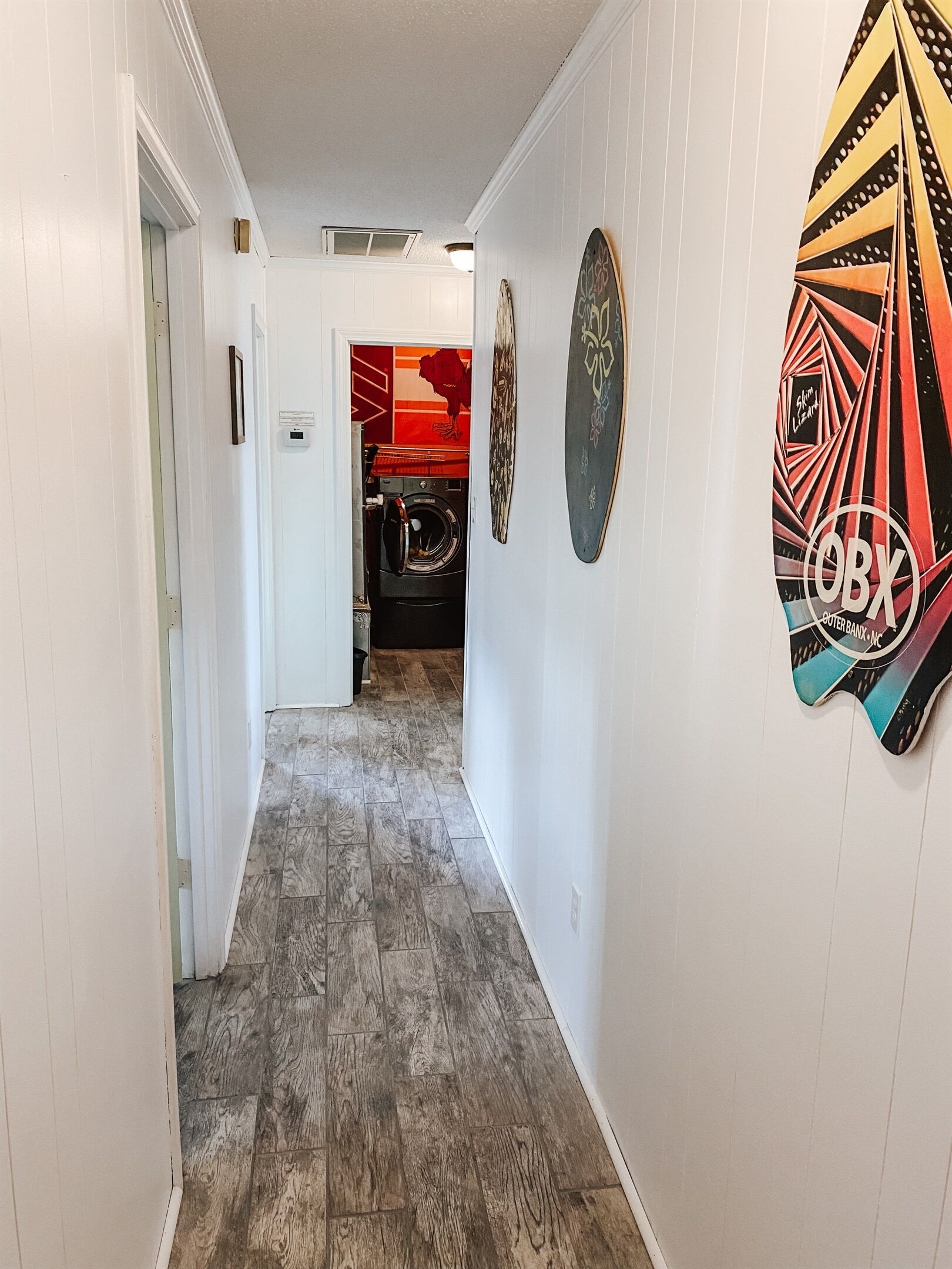 Um corredor com piso de madeira, paredes brancas e uma porta que dá para um quarto com parede vermelha e laranja.