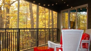 A covered porch with a view of autumn trees, a white wicker chair, and a red table.