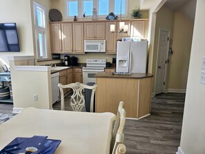 A compact kitchen with wooden cabinets, a microwave, a refrigerator, and a small dining area with a chair.
