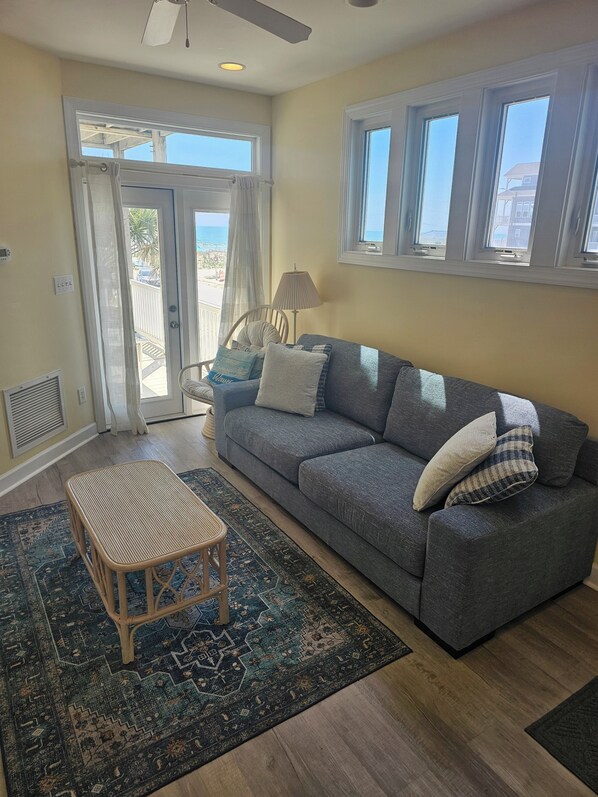 A living room with a gray sofa, a wicker coffee table, a patterned rug, and a view of the beach through a glass door.