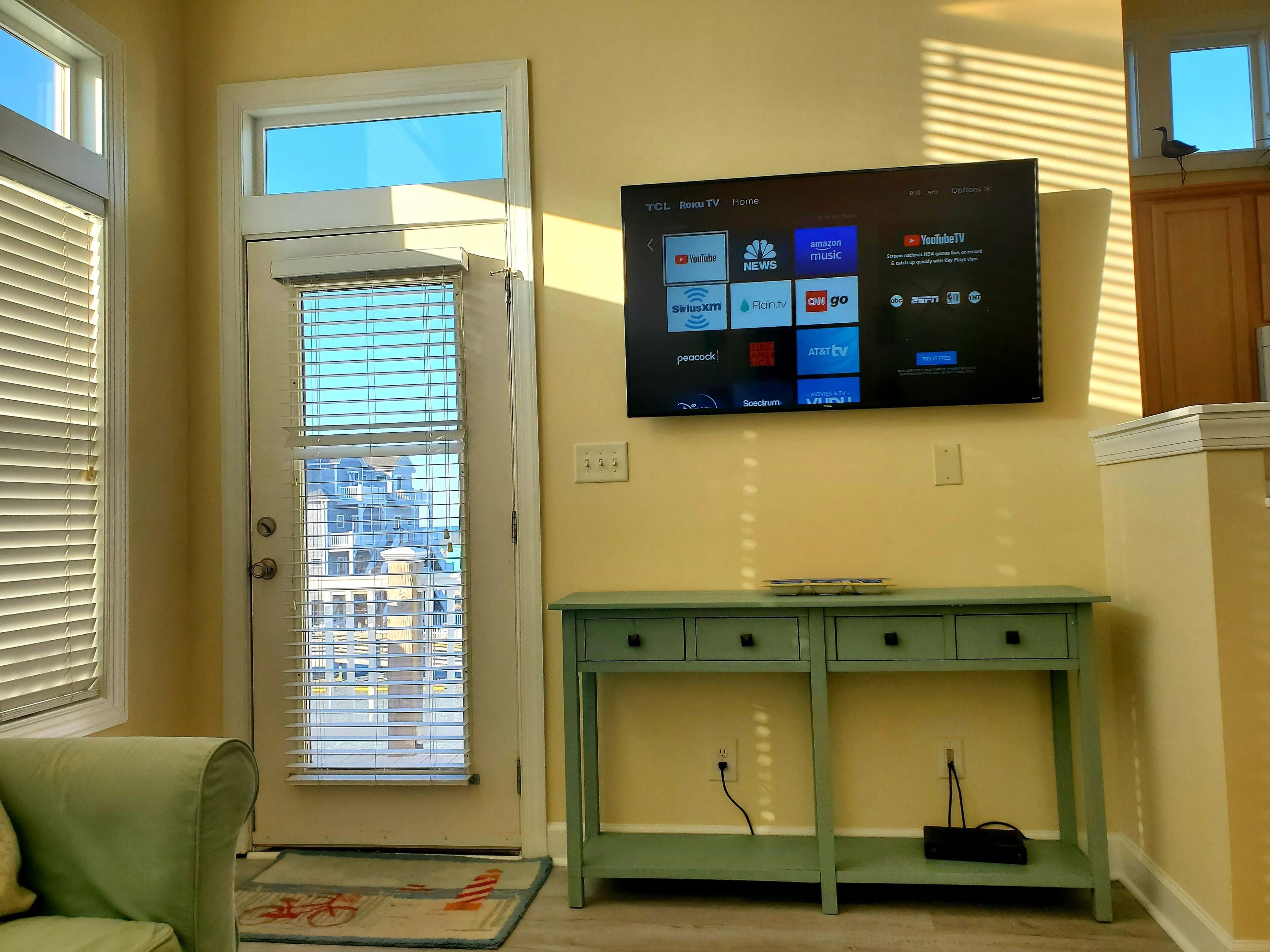 A room with a mounted flat-screen TV displaying various streaming services, a door with a view of buildings outside, a green console table, and a window with blinds.