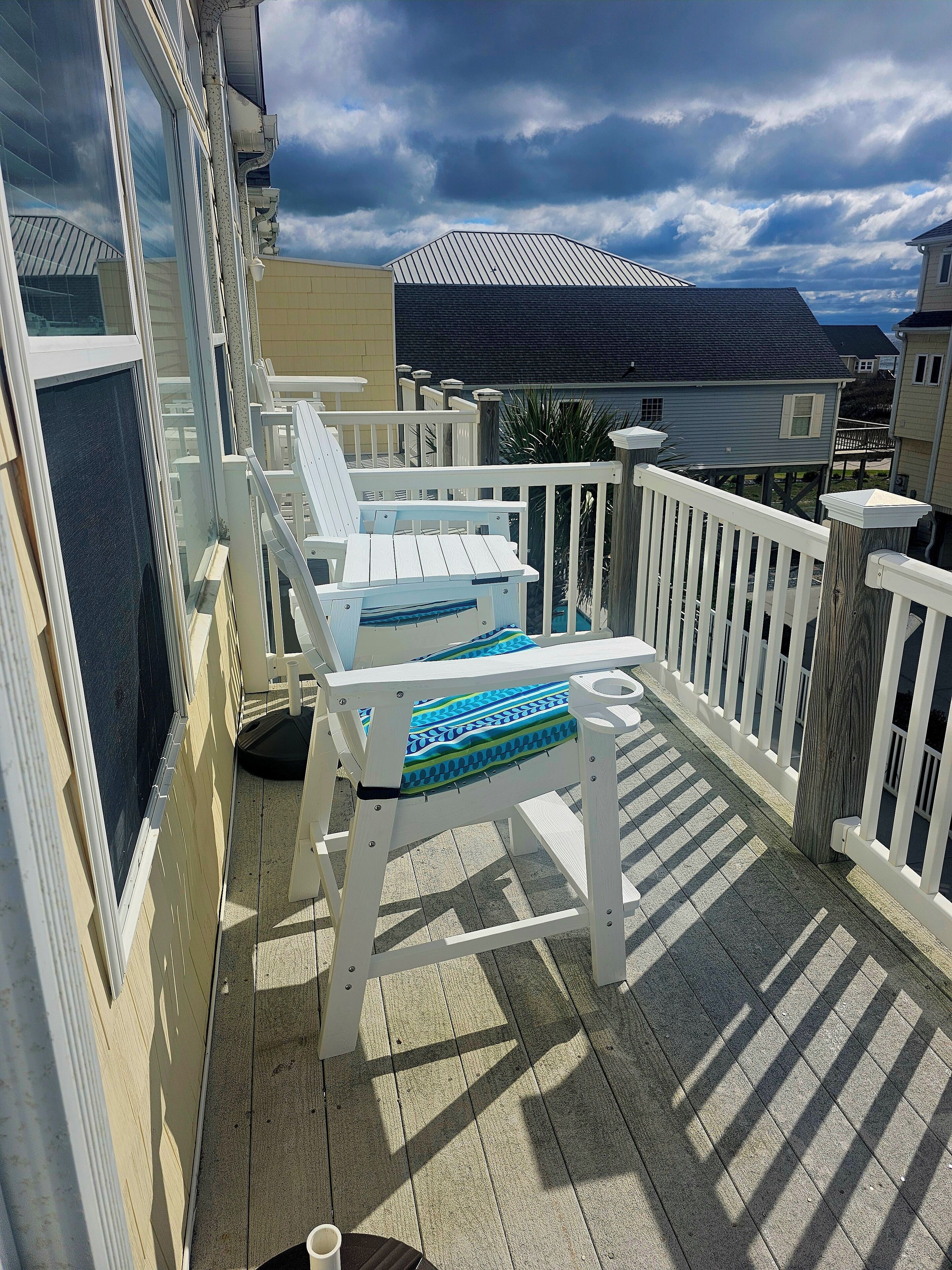 Een houten terras met witte leuningen en twee witte ligstoelen met blauwe kussens.