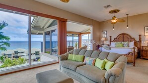 Ein geräumiges Schlafzimmer mit großem Fenster zum Meerblick, ein gemütliches graues Sofa mit grünen Kissen, ein Holzbett mit dekorativer Kopfteil und einem Deckenventilator.