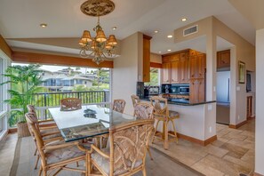 A dining area with a glass table and wicker chairs, a kitchen with wooden cabinets, and a view of the neighborhood. - Kapalua Bay Villa 33B3  Oceanfront Views (Lahaina)