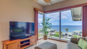 Ein Wohnzimmer mit einem großen Flachbildfernseher auf einer Holzkommode, einem bequemen grauen Sofa und einem großen Fenster mit Blick auf das Meer und Gebäude.