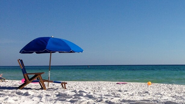 Una playa con una sombrilla azul, una silla de madera y una orilla arenosa.