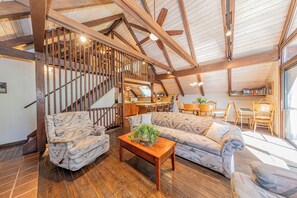 A spacious loft with a high ceiling, wooden beams, a staircase, a living area with patterned sofas, a wooden coffee table, and a kitchen area.