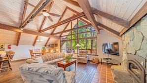 Ein geräumiges Wohnzimmer mit hoher Decke, Holzbalken, einem Kamin aus Stein und großen Fenstern mit Blick auf eine grüne Landschaft.