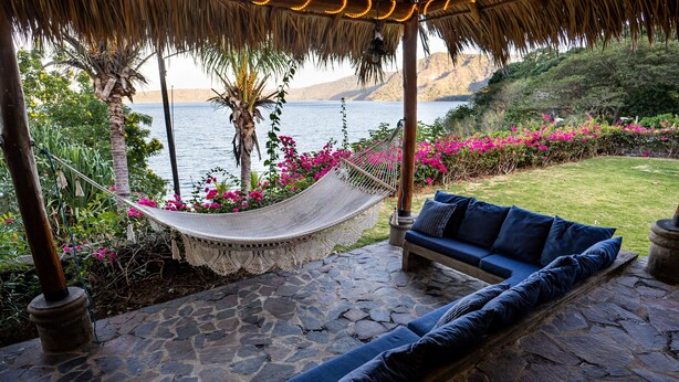 A hammock under a thatched roof, a stone patio, a blue sofa with cushions, and a view of the sea and hills.
