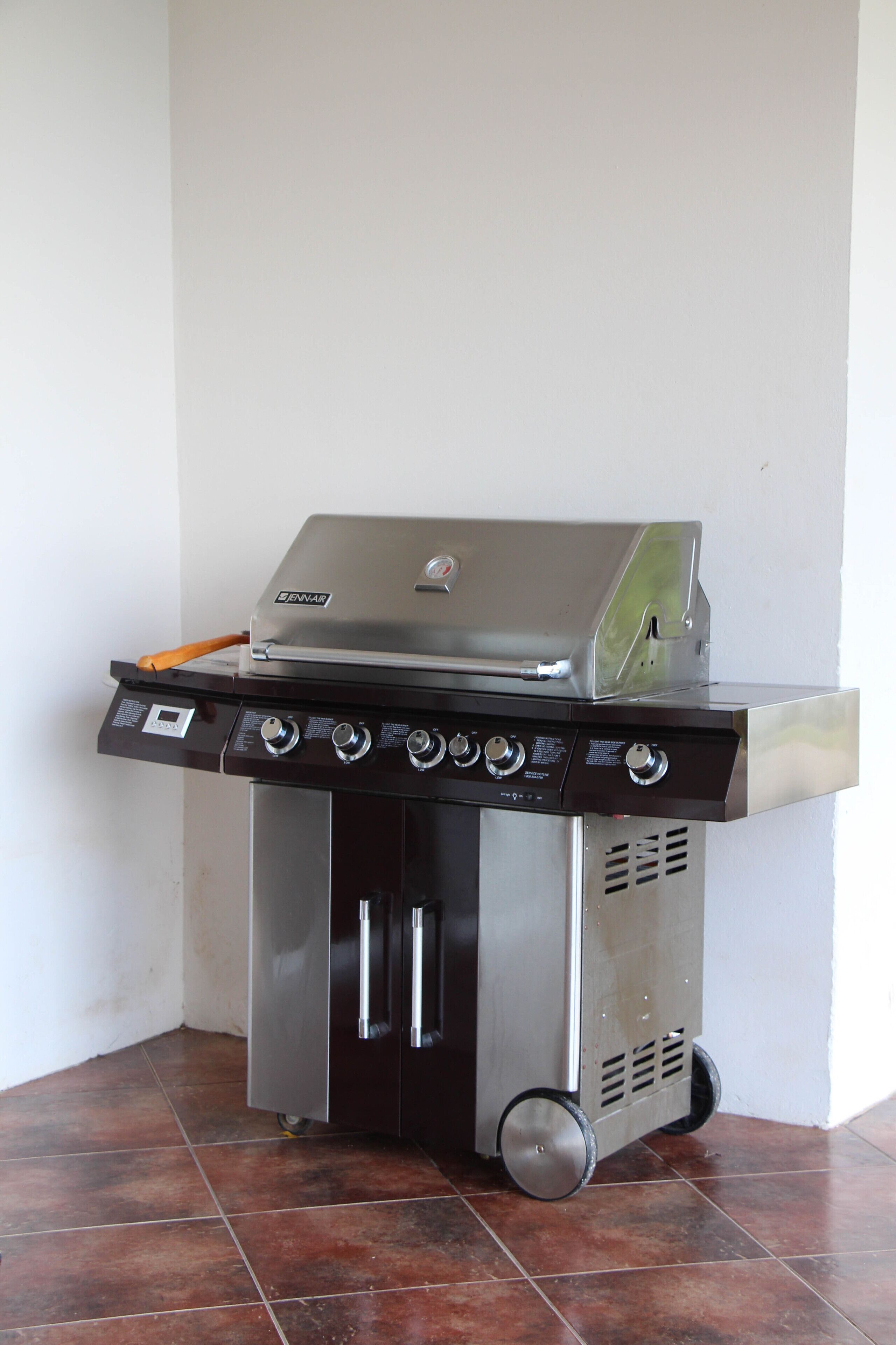 A portable gas grill with a digital display and multiple control knobs, placed on a tiled floor against a plain wall.