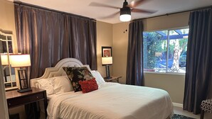 Una cama bien hecha con sábanas blancas, un cojín decorativo y un cabecero. Hay una mesita de noche con una lámpara, y una pequeña mesa con otra lámpara. La habitación tiene un ventilador de techo, una ventana con cortinas y vistas al exterior.