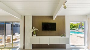 A modern living room with a flat-screen TV mounted on a wooden wall, a white console table, and a glass door leading to an outdoor area with a pool and lounge chairs. - Beautiful! Remodeled Mid-Century Alexander w/ SALTWATER pool, spa, bikes, & more (Palm Springs)