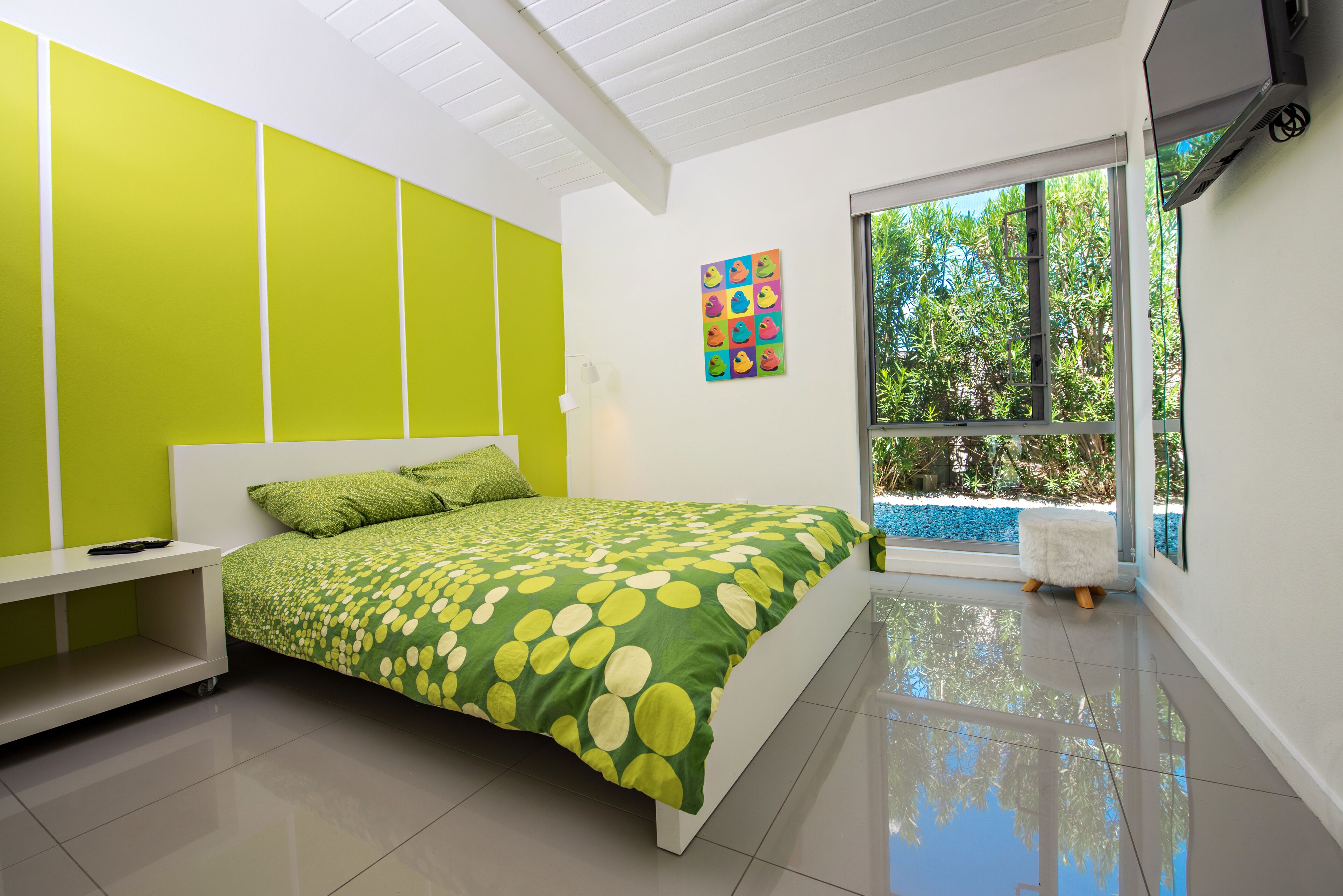 A modern bedroom with a green and white color scheme, a large bed with a green and yellow polka dot bedspread, a white nightstand, and a window with a view of greenery outside.