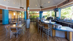 Un restaurant avec des tables et des chaises en bois, un coin bar doté d’un comptoir et de diverses bouteilles, et un ventilateur de plafond.