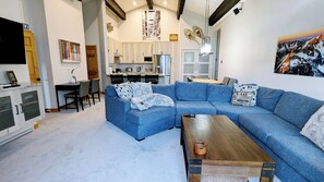 Un salon moderne avec un canapé d’angle bleu, une table basse en bois et une cuisine en arrière-plan.